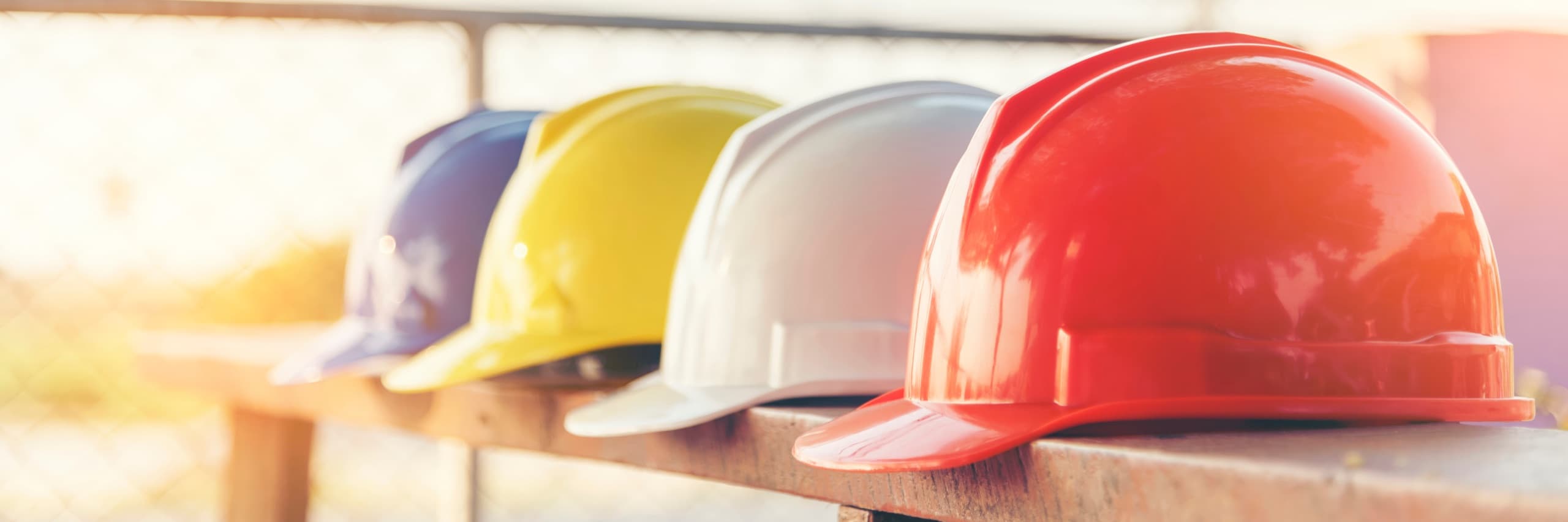 Four clean hard hats in blue, yellow, white and red resting in a row on a timber bench in bright sunlight